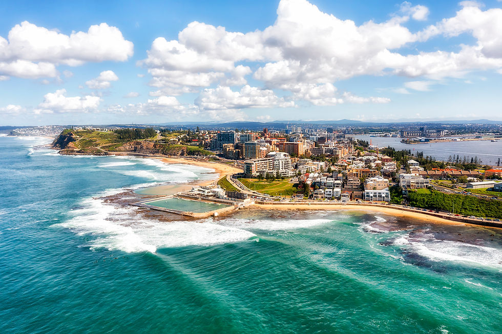 Aerial view of coastal city with clear blue ocean, sandy beaches, lush greenery, and urban skyline under a bright sky with fluffy clouds.
