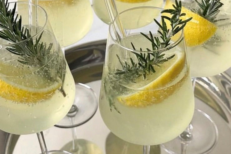 Glasses of sparkling yellow cocktails with lemon slices and rosemary on a silver tray. Bright, refreshing mood.
