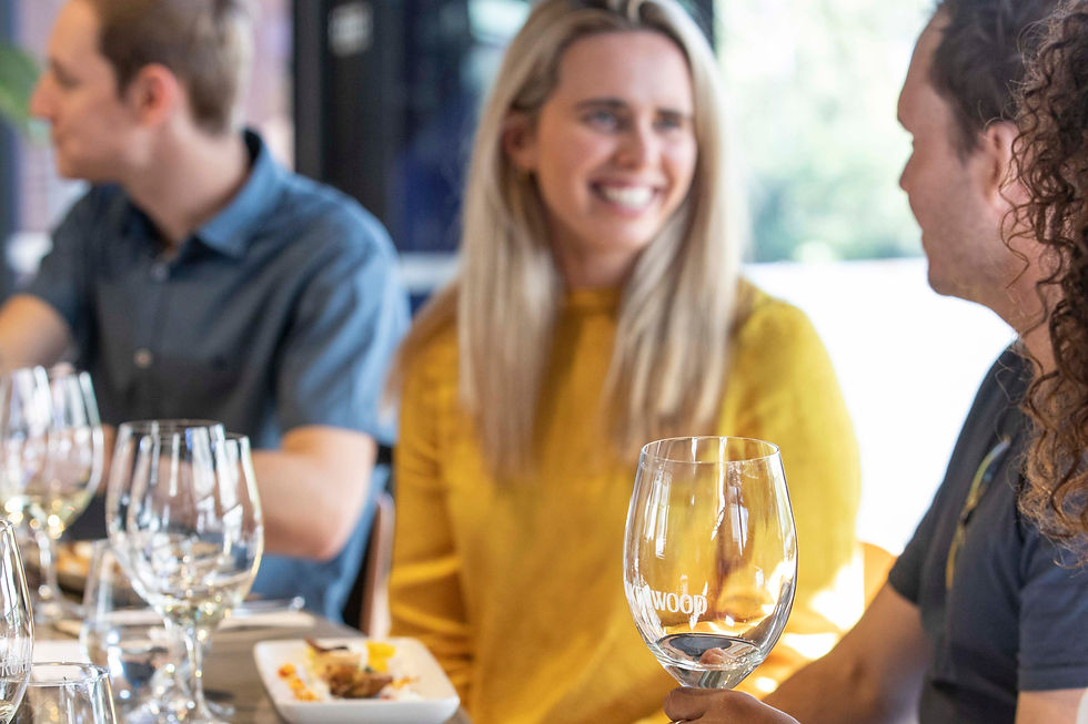 Three people dining, two conversing and laughing, with wine glasses and food on the table. One person wears a yellow shirt. Bright setting.