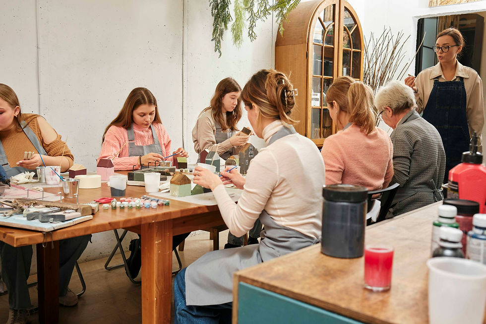People crafting at a wooden table, painting small models. Bright aprons and paint jars visible. Cozy workshop with a relaxed atmosphere.