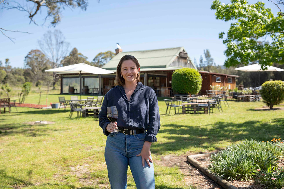 Smiling woman in navy shirt and jeans holds a glass of red wine outside a rustic house with lawn, umbrellas, and patio tables.