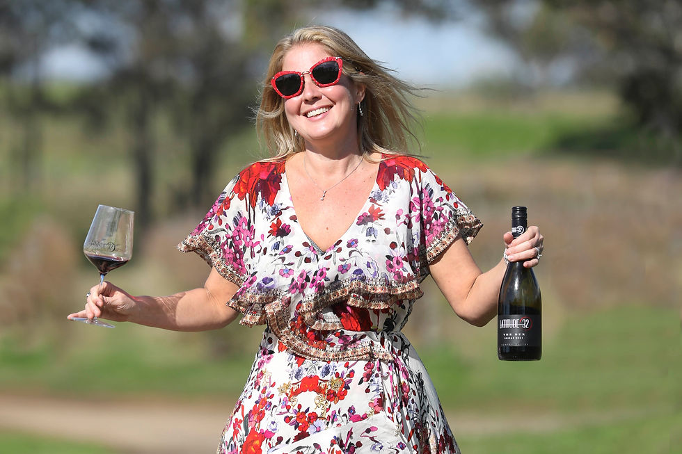 Woman in floral dress and red sunglasses holds wine and glass, smiling in sunny outdoor setting. Wine bottle reads "Latitude 32".
