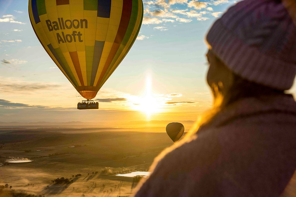 Hot air balloon with "Balloon Aloft" text floats at sunrise. A person in a beanie watches from another balloon. Warm, scenic landscape.