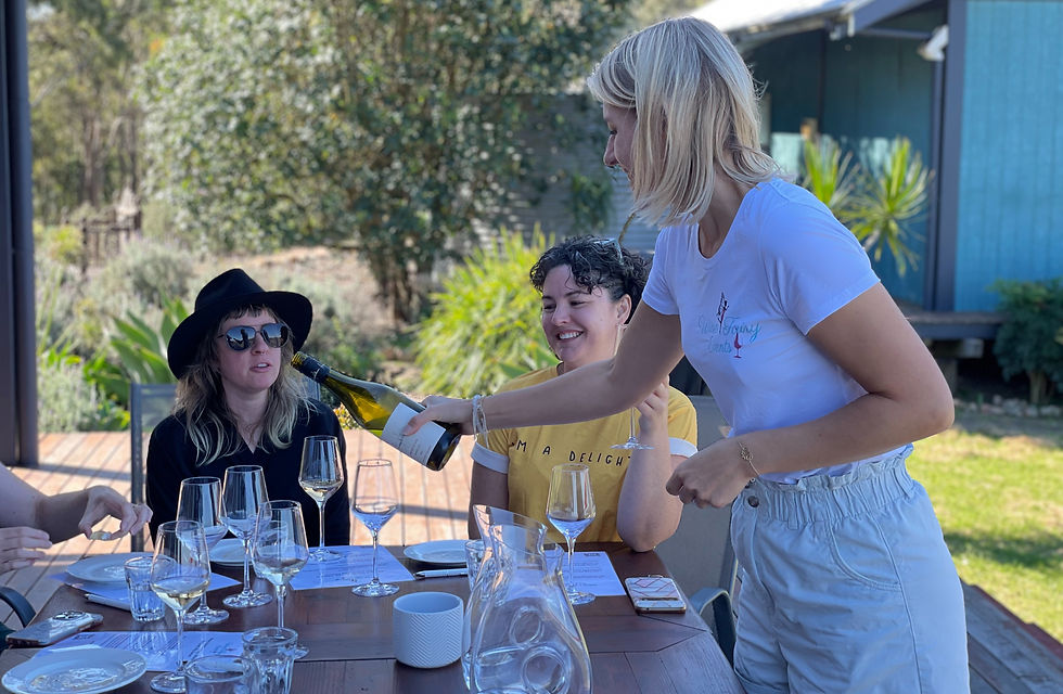 Blonde woman pours white wine for two seated women outdoors. One wears a hat and sunglasses, the other smiles. Sunny day, lush background.