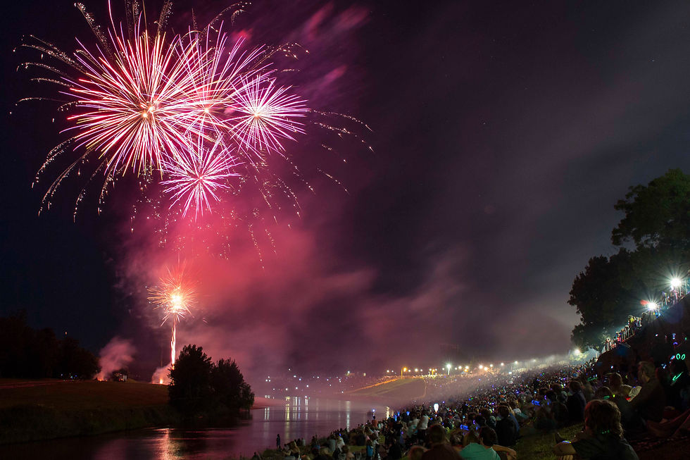 Crowd watches vibrant pink fireworks over a river at night. Smoke fills the sky, lights glow from the audience, creating a festive mood.