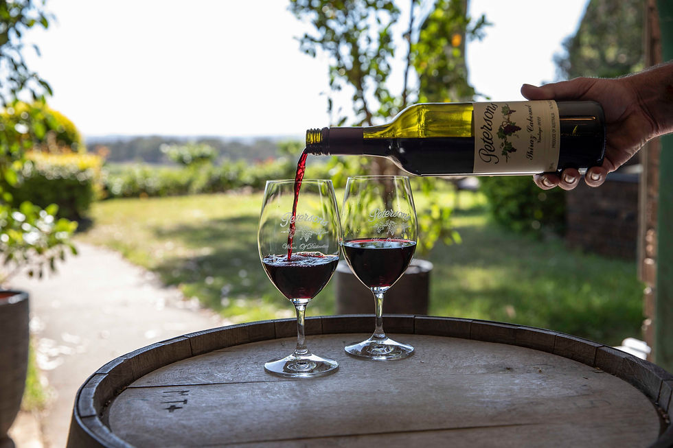 Hand pours red wine from a Petersons bottle into two glasses on a barrel, with a sunny vineyard in the background.