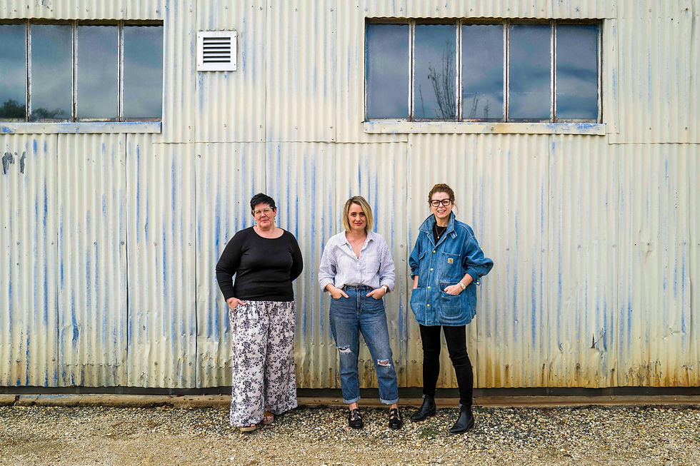 Three women stand smiling in front of a weathered corrugated metal building with large windows.