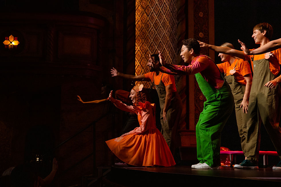 Performers in colorful outfits, including a woman in an orange dress, enthusiastically wave on stage. Ornate wall and dim lighting set the scene.