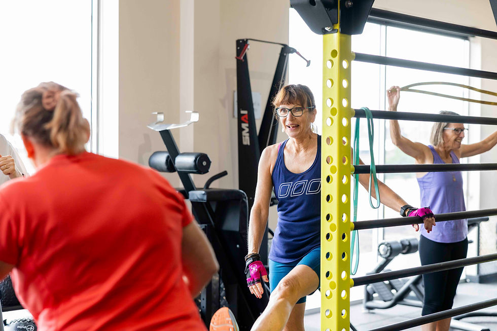 Women exercising in a gym; one stretches near a yellow bar, another uses a resistance band. Bright setting with fitness equipment.