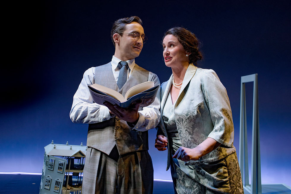 Two people in formal attire stand on stage. One holds a book, both smile, suggesting a lively conversation. A dollhouse is in the background.