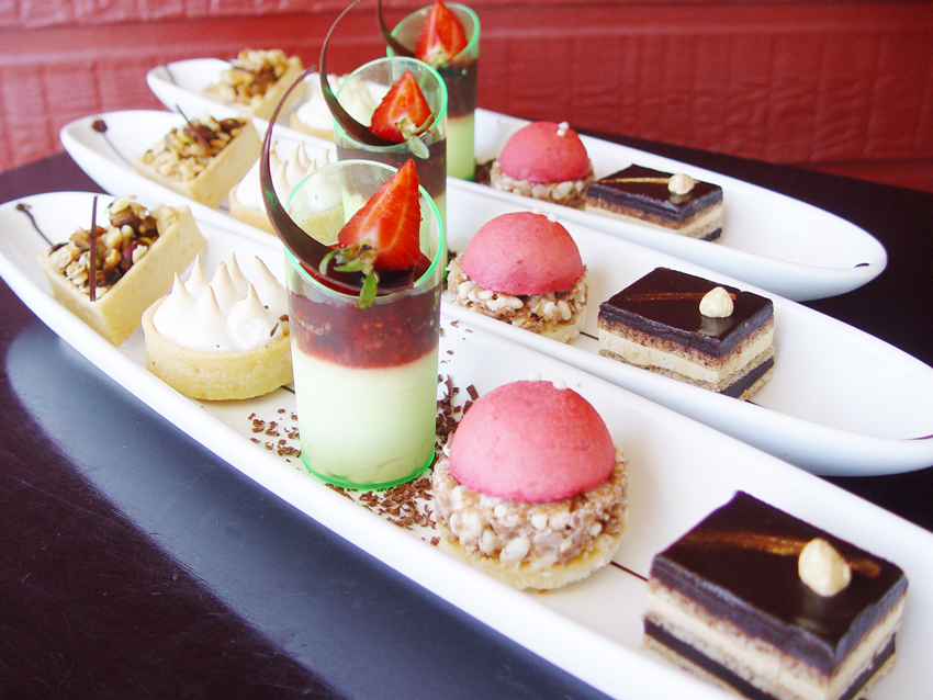 A selection of elegant desserts on white trays: mousse with strawberries, chocolate cake, pink domes, meringue tarts, against a red wall.