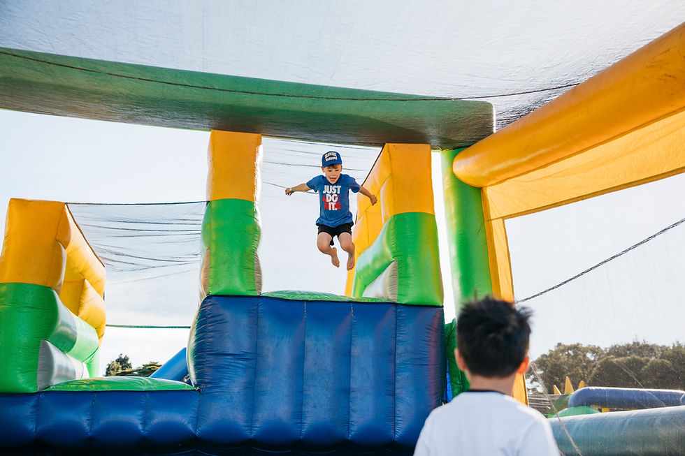 Child in blue shirt and cap jumps on colorful inflatable structure outdoors. Another child watches. Joyful, sunny atmosphere.