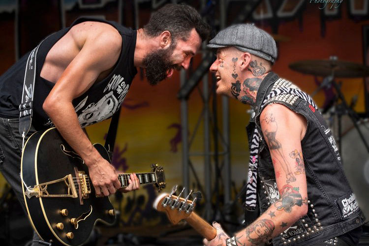 Two tattooed musicians energetically perform with guitars on stage, facing each other. Background has vibrant colors and scaffolding.