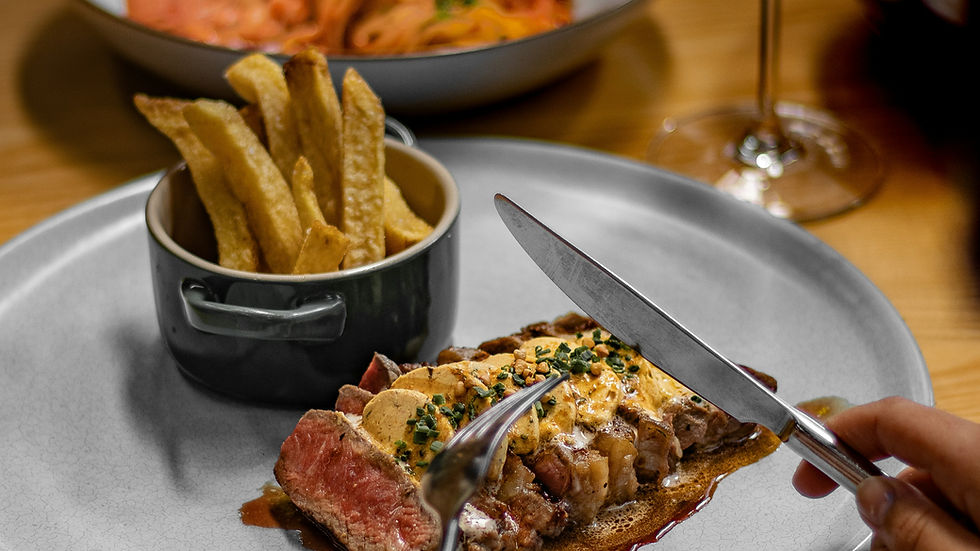 Steak topped with herbs and sauce, being sliced with a knife and fork. Served with fries in a bowl, on a wooden table with dim lighting.