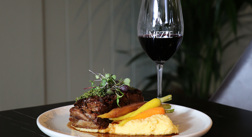 Plated roasted meat with carrots and mashed potatoes beside a red wine glass labeled MEZZE on a dark table.