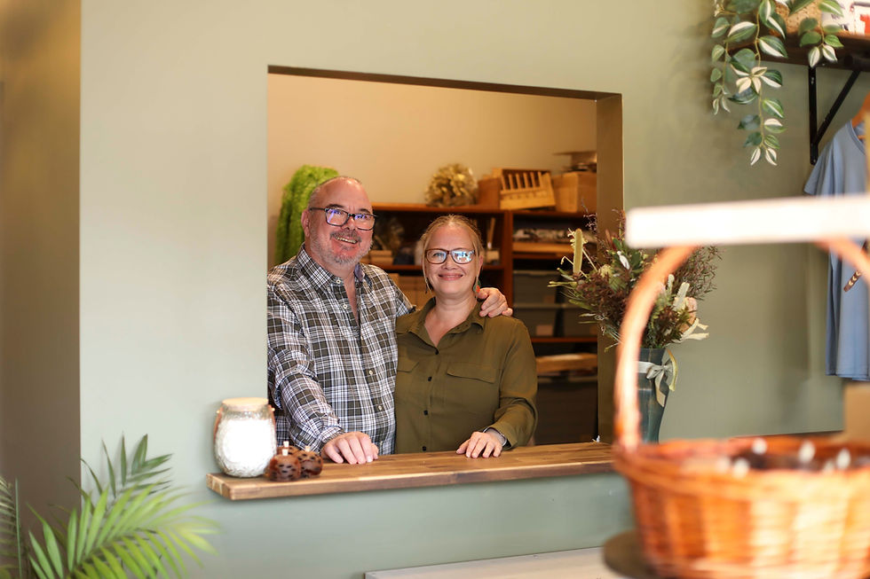 Smiling couple stands at a wooden counter in a cozy shop with green walls, plants, and a wicker basket. Warm, welcoming atmosphere.