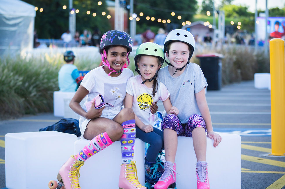 Three kids in helmets sit on a bench, smiling. They're wearing colorful skates and clothing. The background has string lights and greenery.