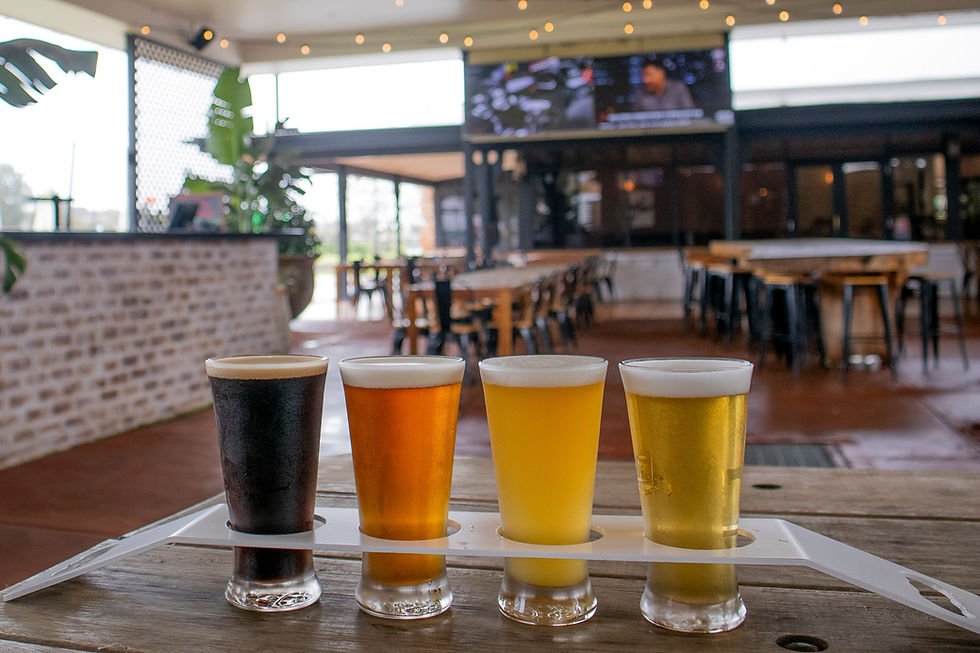 Four beer samples on a wooden table in a cozy bar with brick walls and string lights. Blurred TV playing in the background.