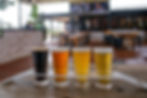 Four beers in a tasting tray on a wooden table. Background shows a rustic bar with brick walls and empty seating. Relaxed atmosphere.