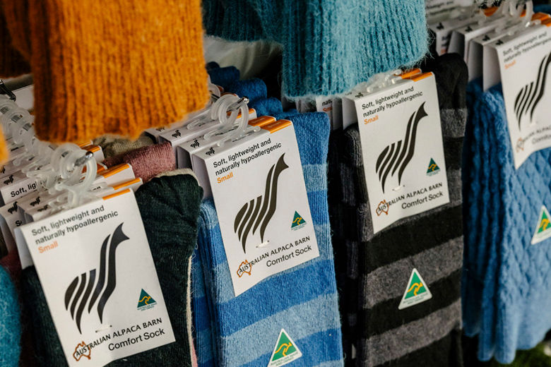 Alpaca socks in various colors (orange, blue, gray) hang on display. Tags read "Australian Alpaca Barn Comfort Sock," highlighting softness.