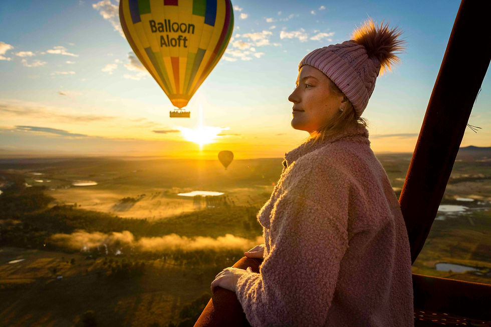 Woman in a pink coat and beanie enjoys sunrise from a hot air balloon. "Balloon Aloft" is visible on another balloon against the sky.