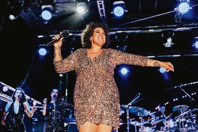 Singer in a sparkling dress performs on stage with a microphone, surrounded by band members and bright stage lights, exuding joy.