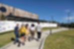 Five people walk toward "BROKENWOOD" winery. Sunny day, blue sky, wooden building, white wall with illustrations, and grassy pathway.