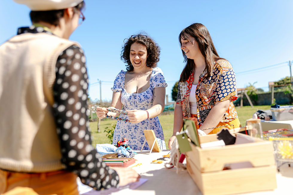 Three people at an outdoor craft fair. Two women in colorful outfits smile while examining handmade items on a table under a clear blue sky.