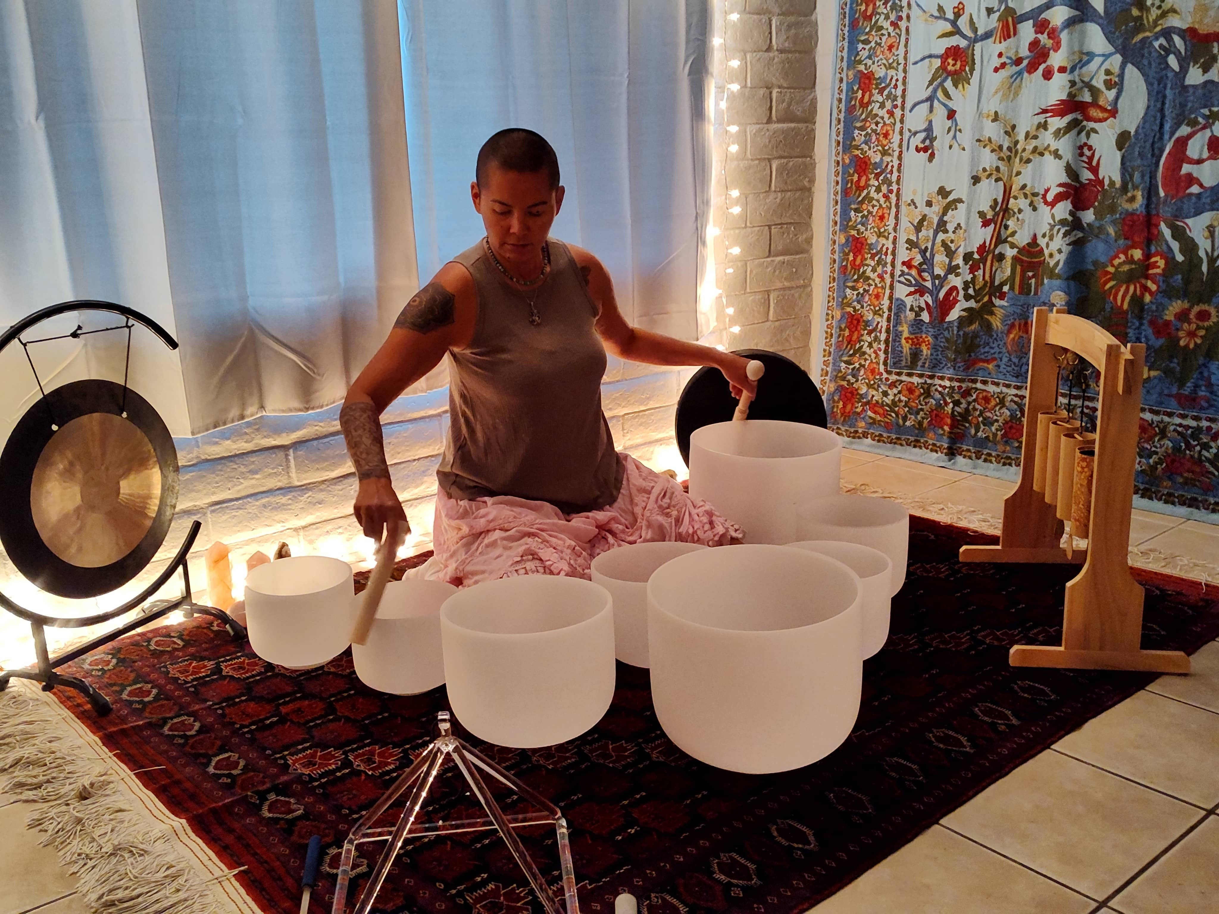A peaceful healing space with singing bowls, gongs and drums set up for a private session. The room features soft lighting, colorful tapestries, and a warm ambiance.