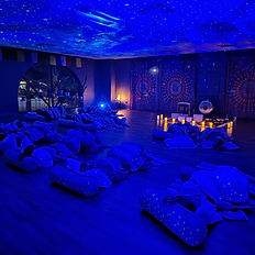 A peaceful healing space with individuals laying down on mats covered by blankets placed near singing bowls. The room features dark lighting, arched windows and a warm ambiance.