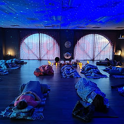A peaceful healing space with individuals laying down on mats covered by blankets placed near singing bowls. The room features dark lighting, arched windows and a warm ambiance.