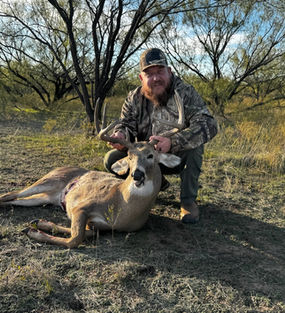 Deer hunter in camouflage kneeling beside a harvested whitetail deer in a grassy field at Cottonwood Ranch.