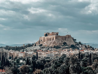 A breathtaking view of the Acropolis of Athens, perched atop a rocky hill, with the Parthenon standing prominently against a backdrop of a partly cloudy sky. Below, the cityscape of Athens stretches out, framed by lush greenery and historic buildings. The foreground features a blurred stone surface, adding depth to the composition.