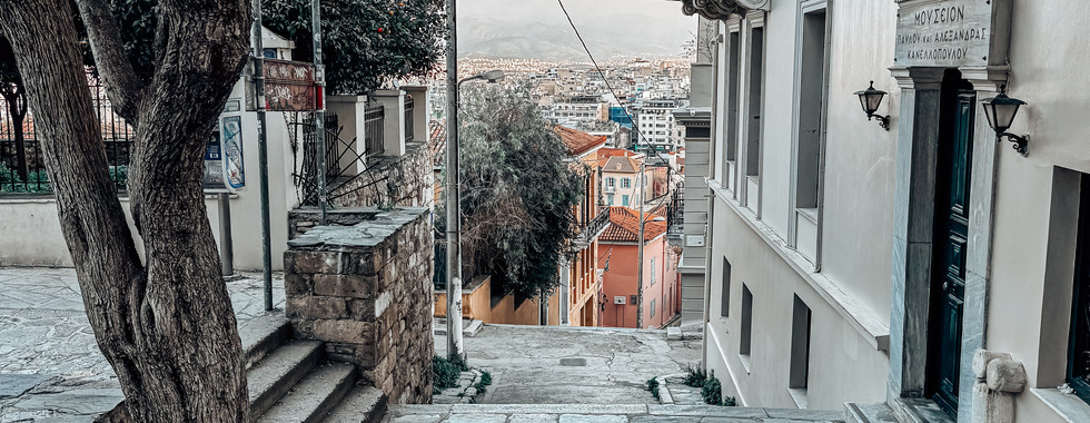 Scenic stone-paved alley in the Anafiotika neighborhood of Athens, Greece, lined with historic white-washed buildings, iron balconies, and a tree with exposed roots, overlooking the city.