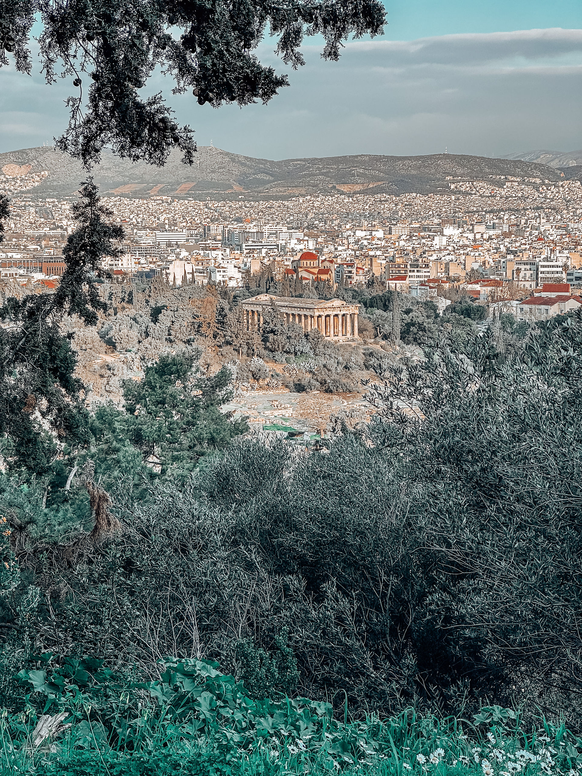 A scenic view of the Temple of Hephaestus in Athens, surrounded by lush greenery, with the cityscape and distant mountains forming a stunning backdrop.