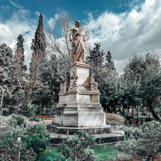 A grand marble statue of Ioannis Kapodistrias, the first Governor of Greece, standing proudly in a lush green park in Athens. The statue portrays him in classical attire, gazing into the distance, while smaller sculpted figures at the base add historical depth. Surrounded by intricate iron fencing, vibrant greenery, and towering cypress trees, the scene captures a blend of history and nature beneath a bright blue sky with scattered clouds.