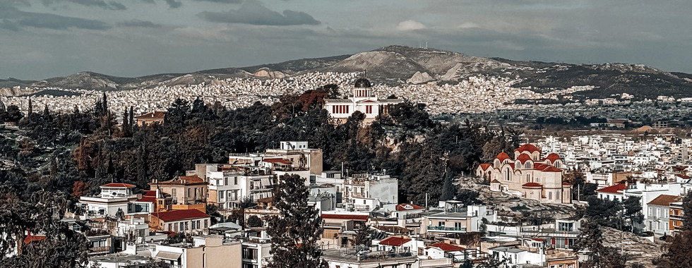 Scenic view of Athens, Greece, from Areopagus Hill, showcasing the Observatory of Athens, red-roofed houses, and distant mountains under a blue sky with scattered clouds.