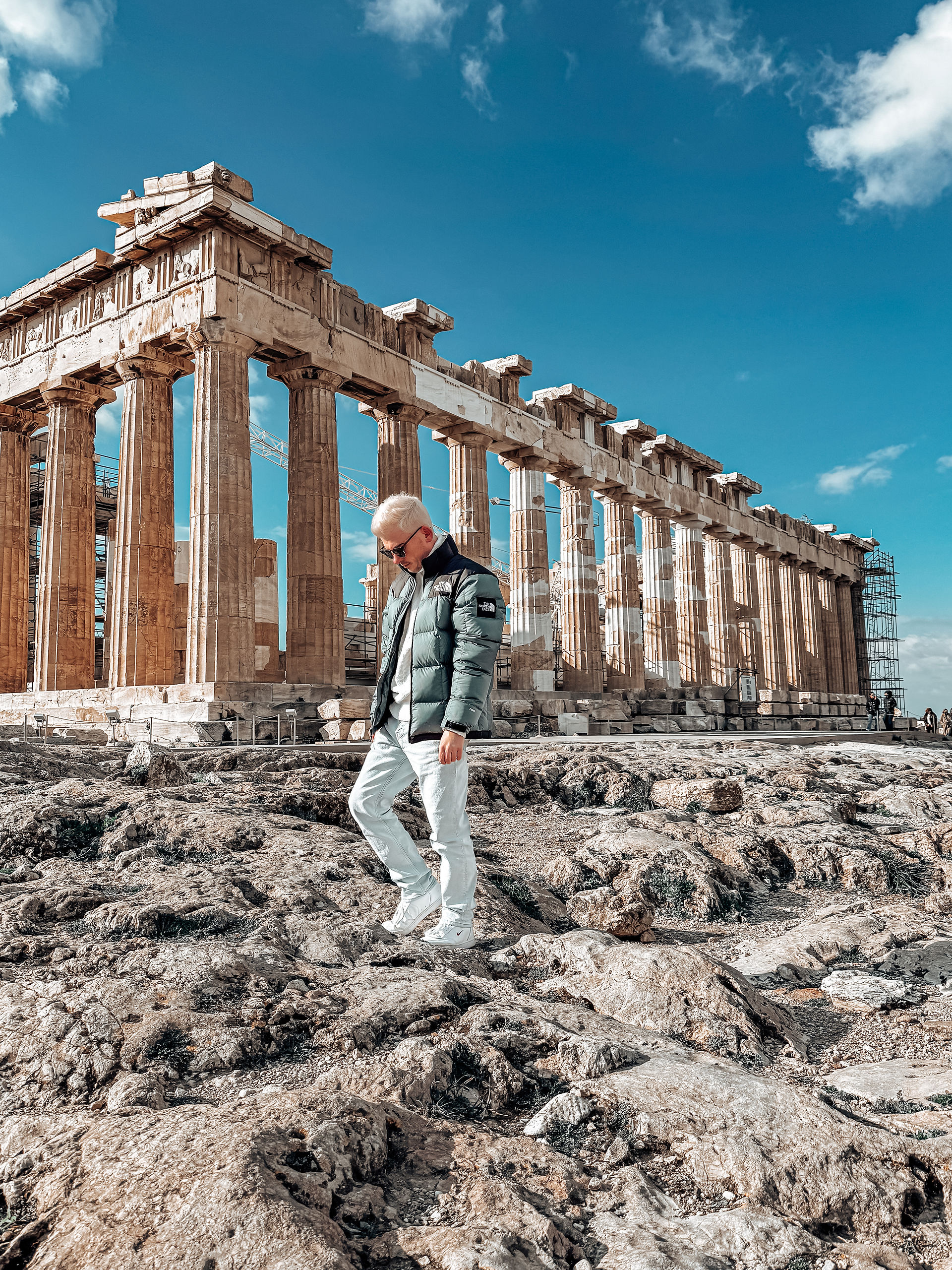 A stylish traveler in a green puffer jacket, white pants, and white sneakers walks across the rugged, rocky terrain of the Acropolis in Athens. The iconic Parthenon, with its grand Doric columns, stands majestically in the background, partially covered with scaffolding for restoration. The deep blue sky with scattered clouds creates a striking contrast against the warm, golden hues of the ancient ruins. The traveler, looking down as he carefully steps over the uneven ground, adds a contemporary touch to this timeless historical setting.