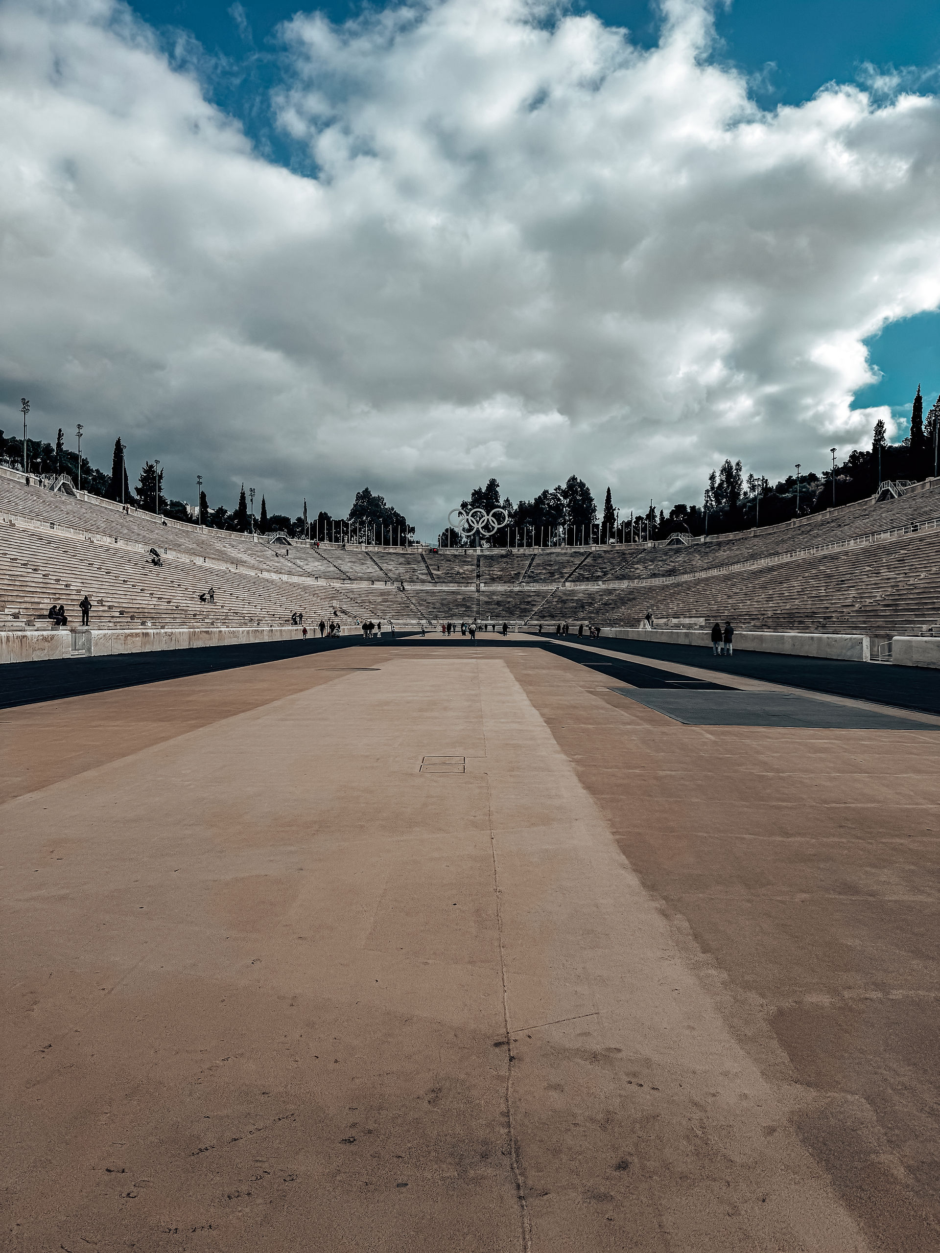 A view of the Panathenaic Stadium in Athens, Greece, taken from the center of the running track. The ancient marble seating curves around both sides, with spectators scattered across the steps. The Olympic rings stand tall at the far end, framed by lush trees. The sky is a mix of bright blue and heavy clouds, casting shadows over the vast open space.