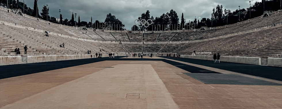 A view of the Panathenaic Stadium in Athens, Greece, taken from the center of the running track. The ancient marble seating curves around both sides, with spectators scattered across the steps. The Olympic rings stand tall at the far end, framed by lush trees. The sky is a mix of bright blue and heavy clouds, casting shadows over the vast open space.