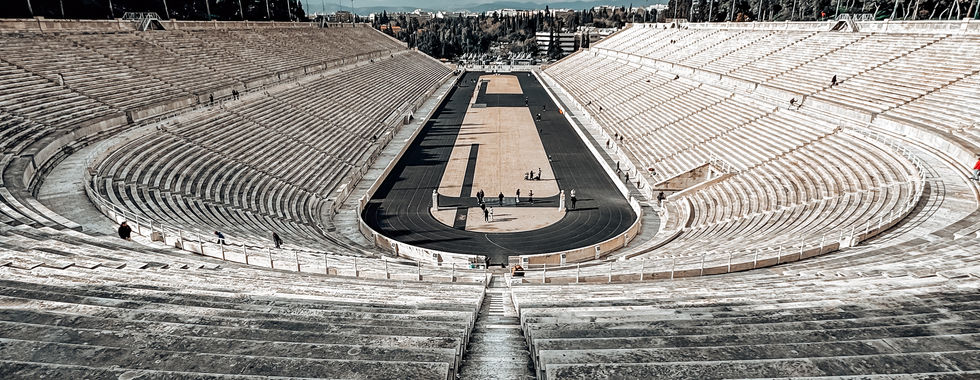 A panoramic view of the Panathenaic Stadium in Athens, Greece, taken from the top of the marble seating area. The long, curved rows of white marble benches extend symmetrically around the central track, which features a black running lane and a sandy competition area. A few visitors can be seen walking on the track, emphasizing the grand scale of the stadium. The background consists of lush green trees, distant city buildings, and a bright blue sky dotted with white clouds.