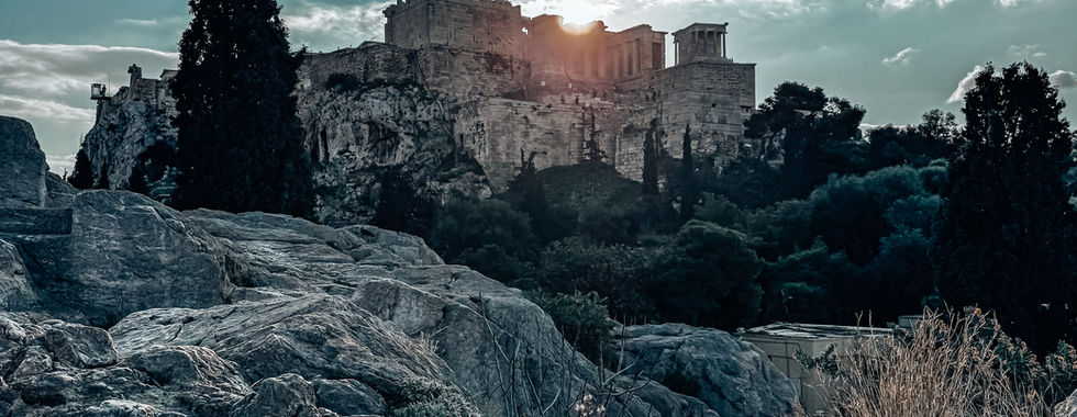 The Acropolis of Athens at sunset, with the Parthenon and Erechtheion temples illuminated by golden light, seen from a rocky viewpoint with dramatic shadows.
