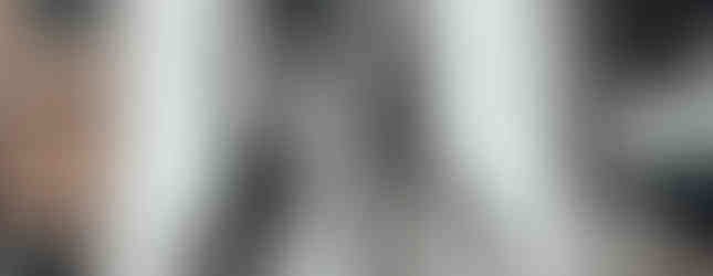 A large-scale black-and-white mural on the side of a building in Athens, Greece, created by the street artist INO. The artwork depicts a young girl with long hair holding a massive sledgehammer, her face partially obscured by shadows. The mural’s monochrome tones contrast with the surrounding urban environment, featuring adjacent buildings, windows, and air conditioning units. A 'PARKING' sign is visible in the lower right corner, adding to the gritty cityscape.