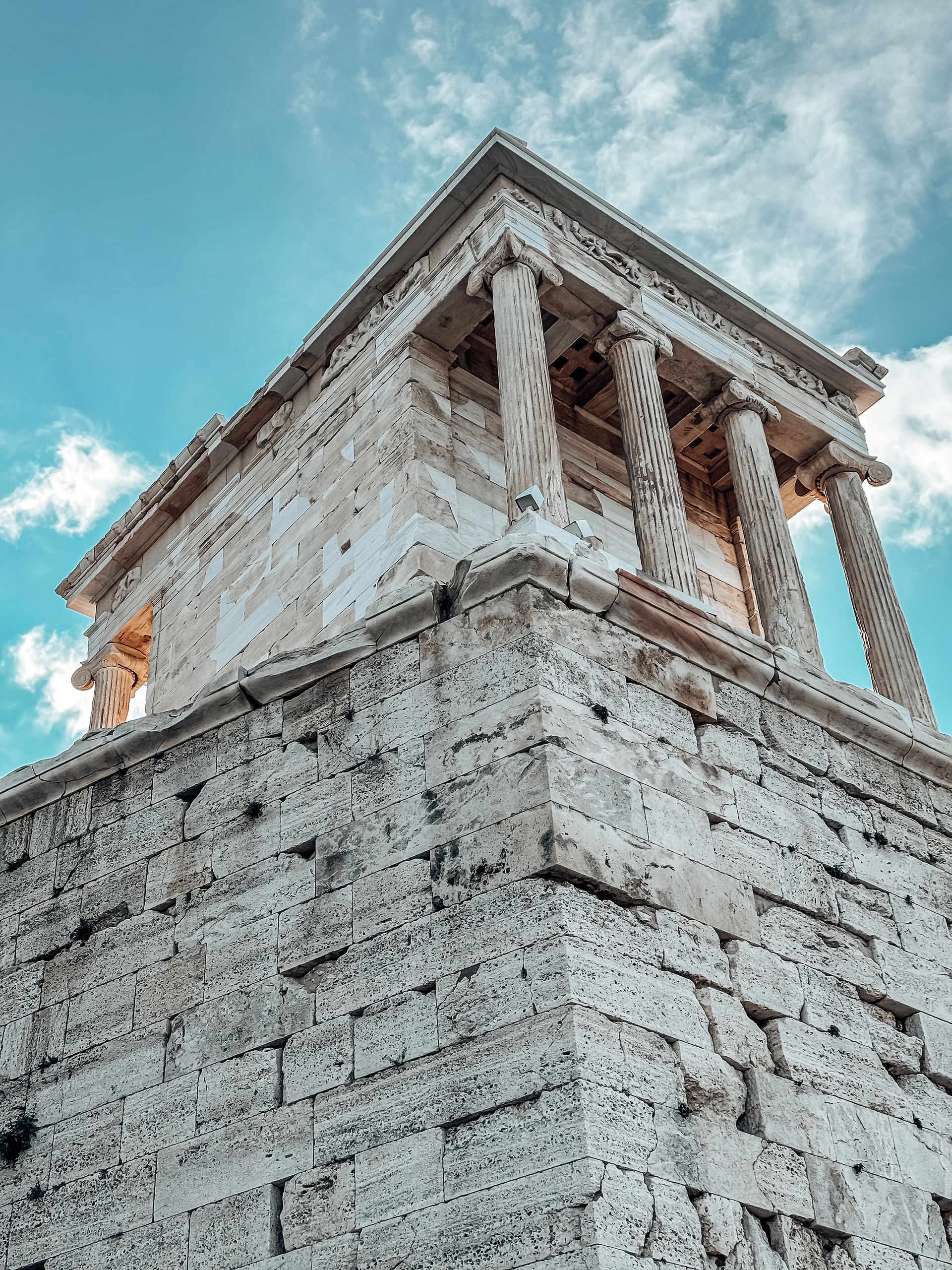 A striking low-angle view of the Temple of Athena Nike perched atop its ancient bastion on the Acropolis of Athens. The temple’s elegant Ionic columns rise gracefully against a backdrop of a vivid blue sky with scattered clouds. The weathered stone foundation, built centuries ago, contrasts beautifully with the restored marble details of the temple, showcasing the harmony between preservation and antiquity. This perspective emphasizes the temple’s historical significance and its enduring presence overlooking the city.
