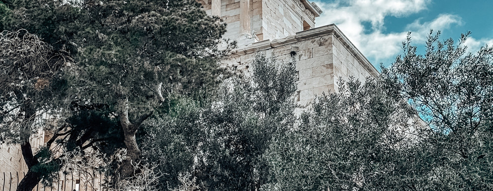 A scenic view of the Temple of Athena Nike, a small yet elegant ancient Greek temple perched on the Acropolis of Athens. The temple, dedicated to the goddess Athena Nike, stands gracefully with its Ionic columns and intricate friezes. The foreground features lush greenery, olive trees, and rugged rocks, adding a natural contrast to the temple’s white marble. The sky above is a mix of soft clouds and blue hues, enhancing the majestic and timeless aura of this historical site.