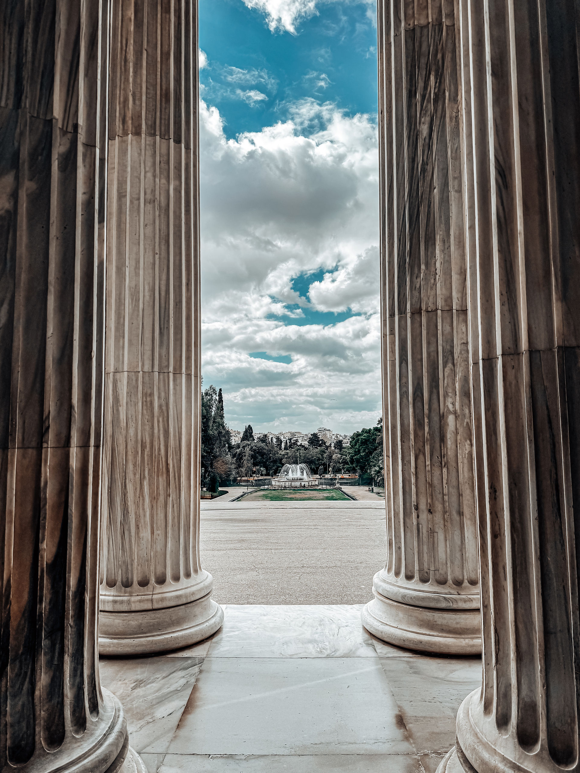 A breathtaking symmetrical view framed by the towering marble columns of the Zappeion Hall in Athens, Greece. Through the grand entrance, a picturesque landscape unfolds, featuring a central fountain, lush greenery, and a cityscape in the distance beneath a sky filled with dramatic clouds.
