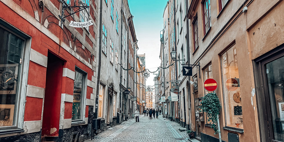 Narrow cobblestone street lined with colorful historic buildings, antique shop signs, people walking, and clear blue sky above.
