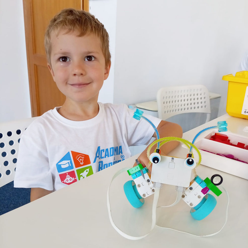 Băiețel zâmbitor în tricou Academia de Robotică, așezat la o masă cu un robot educațional LEGO Education spike essential construit in timpul cursului Robotica Junior 4-6 ani in sala de clasa. 