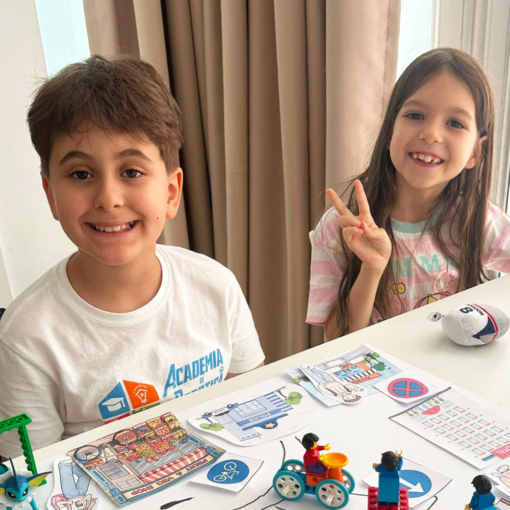 Băiețel zâmbitor în tricou Academia de Robotică, așezat la o masă cu un robot educațional LEGO Education spike essential construit in timpul cursului Robotica Junior 4-6 ani in sala de clasa. 