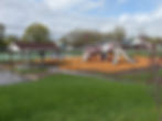 Three grey slides amid other blue, grey and red playground features, such as monkey bars and tunnels, on wood chips surrounded by green grass and a small pavilion.